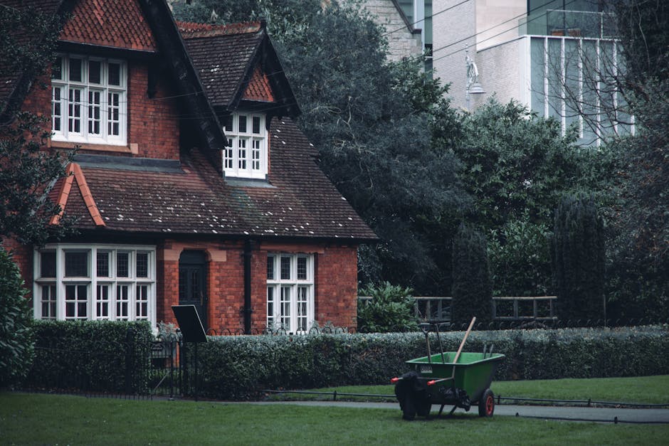 A traditional two-storey red brick house with a steeply pitched tiled roof and white-framed bay windows on the ground and first floors, surrounded by a well-kept garden. The garden features a neatly t