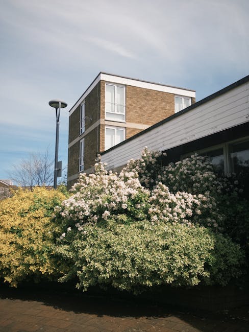 A traditional two-storey red brick house with a steeply pitched tiled roof and white-framed bay windows on the ground and first floors, surrounded by a well-kept garden. The garden features a neatly t