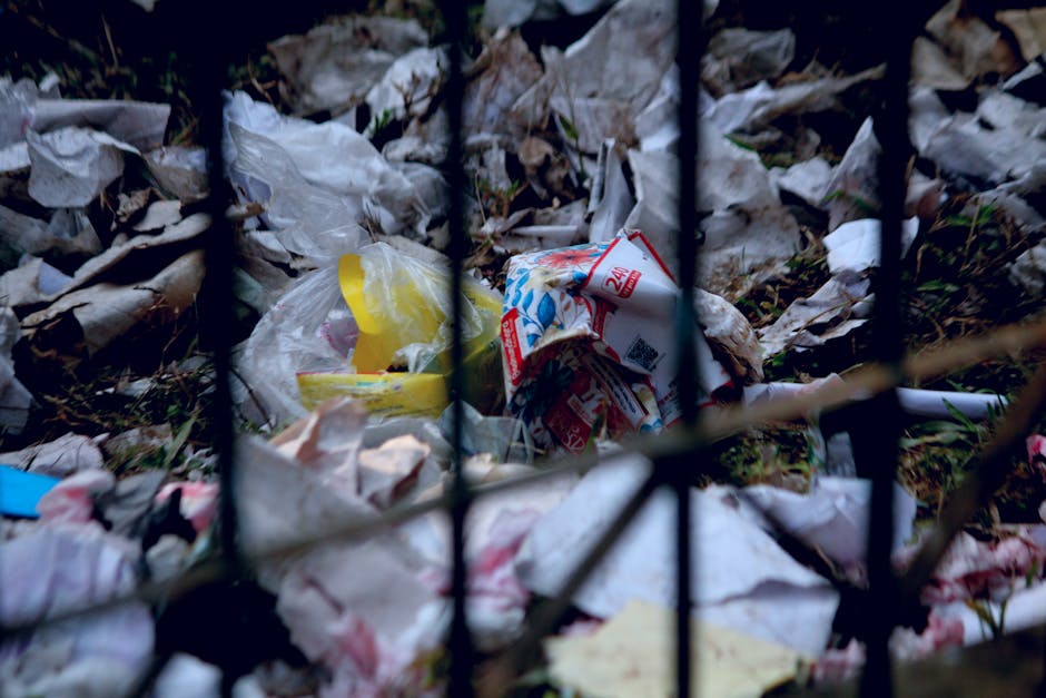 A large, metal wheelie rubbish bin with a slightly rusted and weathered surface, positioned on a concrete pavement against a brick wall and partially surrounded by greenery. The background features a 